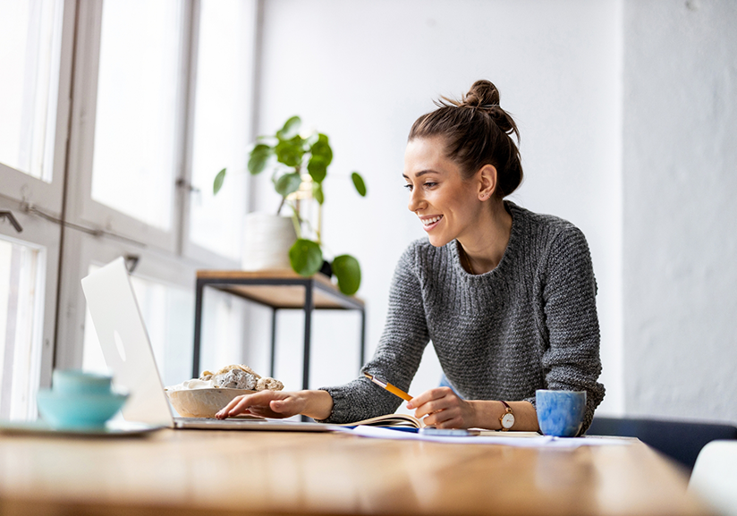 Woman in grey sweater holding pencil and typing on laptop